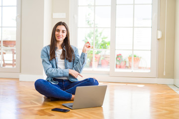 Beautiful young woman sitting on the floor with crossed legs using laptop with a big smile on face, pointing with hand and finger to the side looking at the camera.