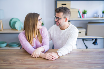 Young beautiful couple sitting on the table at home, hugging in love very happy for moving to new home with cardboard boxes behind them