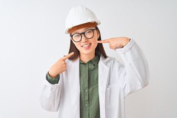 Young chinese engineer woman wearing coat helmet glasses over isolated white background smiling cheerful showing and pointing with fingers teeth and mouth. Dental health concept.