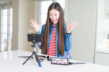 Beautiful Asian influencer woman recording make up tutorial using smartphone camera celebrating crazy and amazed for success with arms raised and open eyes screaming excited. Winner concept