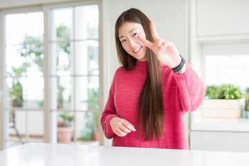 Beautiful Asian woman wearing pink sweater on white table smiling looking to the camera showing fingers doing victory sign. Number two.