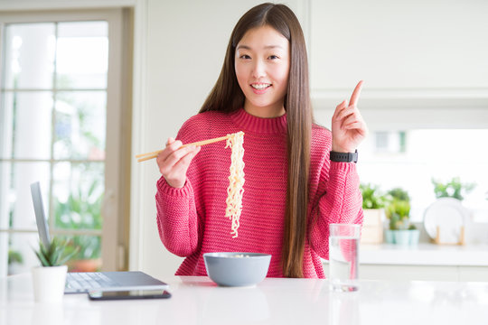 Beautiful Asian woman working using laptop and eating asian noodles surprised with an idea or question pointing finger with happy face, number one