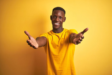 Young african american man wearing casual t-shirt standing over isolated yellow background looking at the camera smiling with open arms for hug. Cheerful expression embracing happiness. - obrazy, fototapety, plakaty