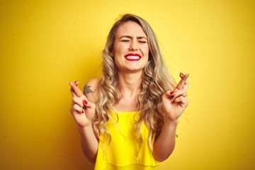 Young attactive woman wearing t-shirt standing over yellow isolated background gesturing finger crossed smiling with hope and eyes closed. Luck and superstitious concept.