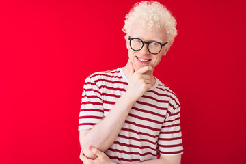 Young albino blond man wearing striped t-shirt and glasses over isolated red background looking...