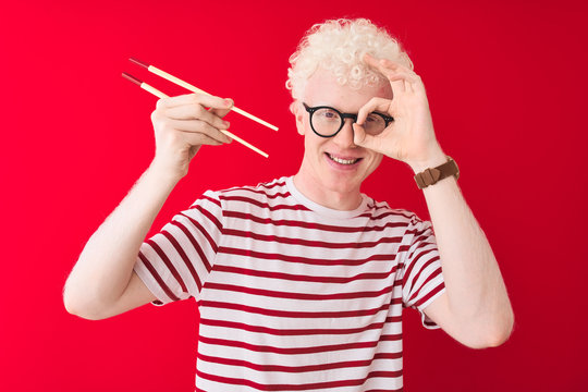 Young albino blond man holding chopsticks standing over isolated white background with happy face smiling doing ok sign with hand on eye looking through fingers