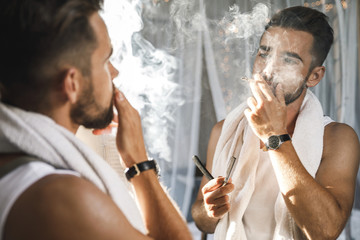 Man looking in the mirror, smoking a cigarette and shaving his beard with a straight razor