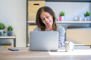 Middle age senior woman sitting at the table at home working using computer laptop sticking tongue out happy with funny expression. Emotion concept.