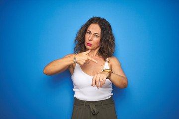 Middle age senior woman with curly hair standing over blue isolated background In hurry pointing to...