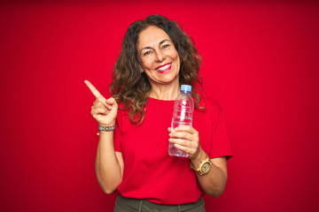 Middle age senior woman holding plastic water bottle over red isolated background very happy pointing with hand and finger to the side