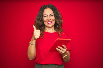 Middle age senior woman using touchpad tablet over red isolated background happy with big smile doing ok sign, thumb up with fingers, excellent sign