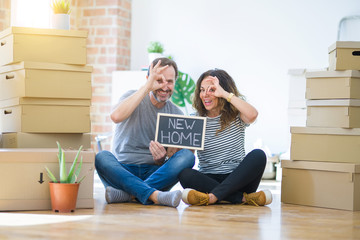 Middle age senior couple sitting on the floor holding blackboard moving to a new home with happy face smiling doing ok sign with hand on eye looking through fingers