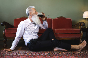Handsome bearded senior man sitting on the floor and drinking red wine