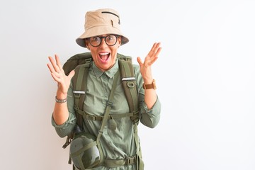 Fototapeta premium Middle age hiker woman wearing backpack canteen hat glasses over isolated white background celebrating crazy and amazed for success with arms raised and open eyes screaming excited. Winner concept
