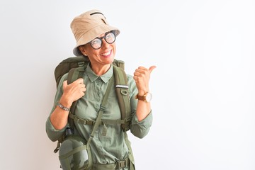 Fototapeta premium Middle age hiker woman wearing backpack canteen hat glasses over isolated white background smiling with happy face looking and pointing to the side with thumb up.