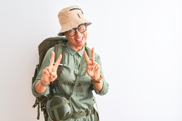 Fototapeta premium Middle age hiker woman wearing backpack canteen hat glasses over isolated white background smiling looking to the camera showing fingers doing victory sign. Number two.