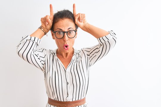 Middle age businesswoman wearing striped dress and glasses over isolated white background doing funny gesture with finger over head as bull horns