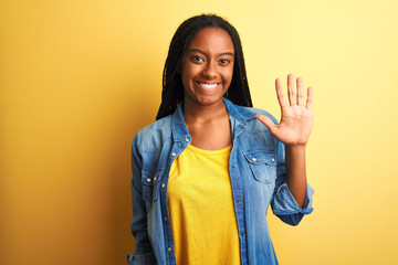 Young african american woman wearing denim shirt standing over isolated yellow background showing and pointing up with fingers number five while smiling confident and happy.