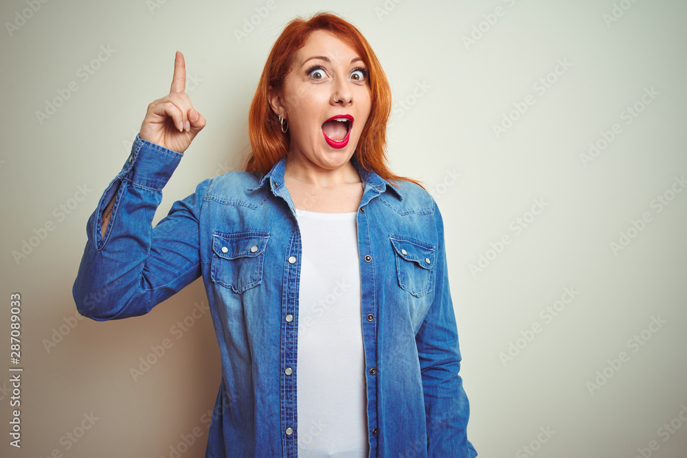 Poster Young beautiful redhead woman wearing denim shirt standing over white isolated background pointing finger up with successful idea. Exited and happy. Number one.