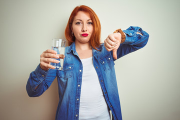 Young beautiful redhead woman drinking glass of water over white isolated background with angry...