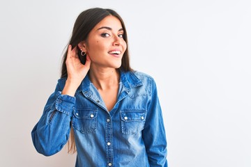 Young beautiful woman wearing casual denim shirt standing over isolated white background smiling with hand over ear listening an hearing to rumor or gossip. Deafness concept.