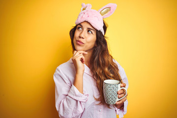 Young woman wearing pajama and mask drinking coffee over yellow isolated background serious face...