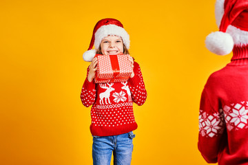 Cheerful kids in Santa hats and red sweaters standing with presents in gift boxes on yellow background