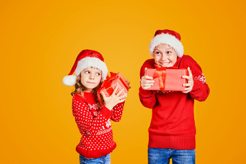 Cheerful kids in Santa hats and red sweaters standing with presents checking what is inside of gift box on yellow background