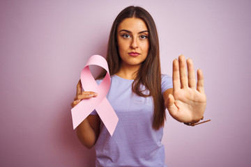 Young beautiful woman holding cancer ribbon standing over isolated pink background with open hand...
