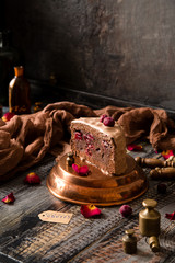 homemade chocolate biscuit cake with chocolate creamy frosting and cherries inside and on top stands on copper pan on wooden table with brown napkin opposite concrete wall