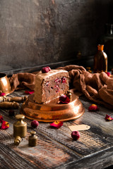 homemade chocolate biscuit cake with chocolate creamy frosting and cherries inside and on top stands on copper pan on wooden table with brown napkin opposite concrete wall