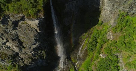 Aerial: Rainbow appearing over misty waterfall falling from rocky cliff on moss covered land during sunny day - Geiranger Fjord, Norway
