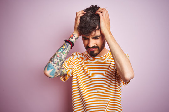Young man with tattoo wearing yellow striped t-shirt standing over isolated pink background suffering from headache desperate and stressed because pain and migraine. Hands on head.