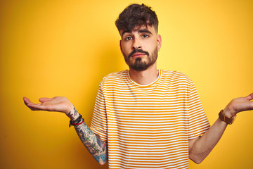 Young man with tattoo wearing striped t-shirt standing over isolated yellow background clueless and...