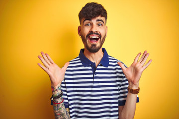 Young man with tattoo wearing striped polo standing over isolated yellow background celebrating crazy and amazed for success with arms raised and open eyes screaming excited. Winner concept