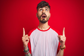 Young man with tattoo wearing striped t-shirt standing over isolated red background amazed and surprised looking up and pointing with fingers and raised arms.