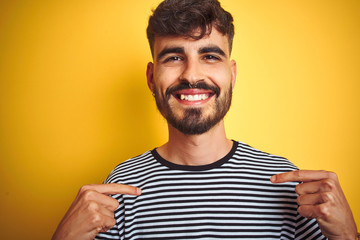 Young man with tattoo wearing striped t-shirt standing over isolated yellow background looking confident with smile on face, pointing oneself with fingers proud and happy.