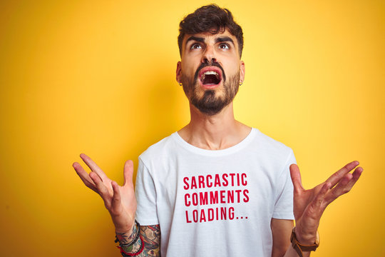 Young man with tattoo wearing fanny t-shirt standing over isolated yellow background crazy and mad shouting and yelling with aggressive expression and arms raised. Frustration concept.