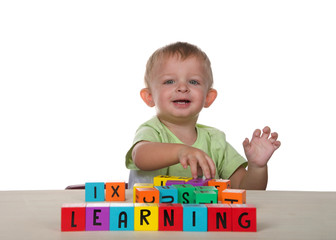 Caucasian toddler male child playing learning with colorful wooden lettered blocks on light wood table, looking towards viewer with happy expression. Isolated on white background.