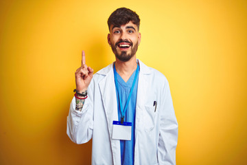 Young doctor man with tattoo wearing id card standing over isolated yellow background pointing finger up with successful idea. Exited and happy. Number one.