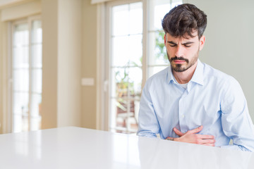 Young businesss man sitting on white table with hand on stomach because nausea, painful disease feeling unwell. Ache concept.