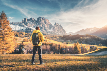 Young man with backpack standing on the hill against the mountains at sunset in autumn. Landscape with sporty guy, meadow,  snowy rocks, orange trees, houses, blue sky. Travel in Italy in fall