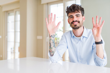Young businesss man sitting on white table showing and pointing up with fingers number nine while smiling confident and happy.