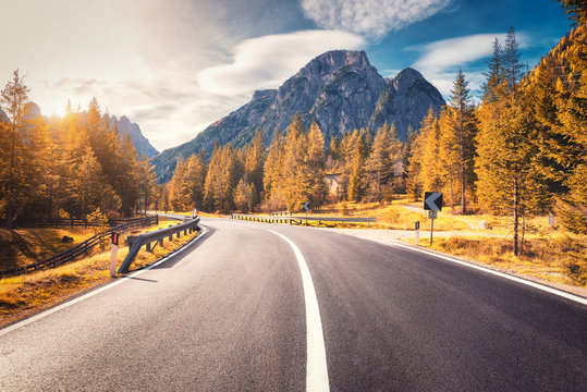 Road in autumn forest at sunset in Italy. Beautiful mountain roadway, trees with orange foliage and sunlight. Landscape with empty asphalt road through woodland, blue sky, high rocks in fall. Travel