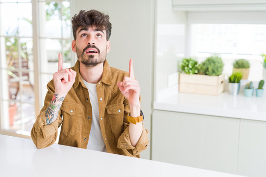 Young man wearing casual jacket sitting on white table amazed and surprised looking up and pointing with fingers and raised arms.