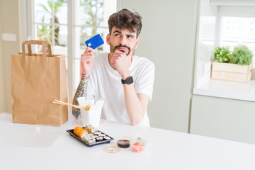 Young man eating asian sushi from home delivery using credit card as payment serious face thinking about question, very confused idea
