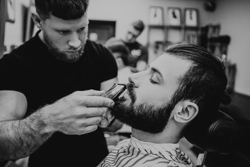 Getting perfect shape. Close-up side view of young stylish bearded man getting beard haircut by bearded, muscular hairdresser or barber at barbershop.Selective focus, noise effect