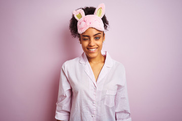 Young african american woman wearing pajama and mask over isolated pink background winking looking...