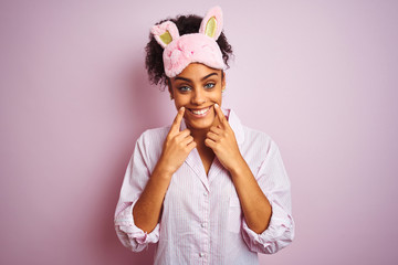 Young african american woman wearing pajama and mask over isolated pink background Smiling with open mouth, fingers pointing and forcing cheerful smile