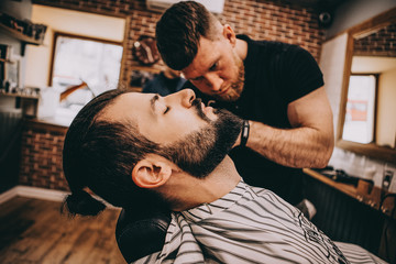 Getting perfect shape. Close-up side view of young stylish bearded man getting beard haircut by...